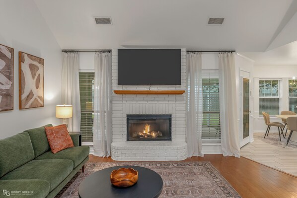 A living room with a fireplace, a green sofa, a black coffee table, and a flat-screen TV mounted above the fireplace.