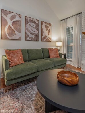 A green sofa with patterned cushions, a round coffee table, and abstract artwork on the wall. - Modern Ranch with King Bed + Spa Bath + Patio + Easy Access (Bossier City)