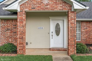 A brick house with a white garage door, a mailbox, and a small front yard with grass and bushes. - Modern Ranch with King Bed + Spa Bath + Patio + Easy Access (Bossier City)