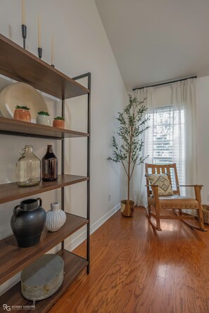 A room with wooden flooring, a bookshelf with decorative items, a potted plant, a wooden chair, and a window with curtains. - Modern Ranch with King Bed + Spa Bath + Patio + Easy Access (Bossier City)