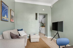 Ein Wohnzimmer mit einer Couch, einem Hocker und einem Fernseher auf einem Schrank. Im Hintergrund ist eine Küche erkennbar.