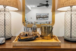 Ein Zimmer mit einem Holftablett, auf dem ein Cocktail-Shaker, eine Flasche und ein Flaschenöffner stehen. Es gibt einen Spiegel mit goldenem Rahmen, einen Deckenventilator und ein Bild an der Wand.