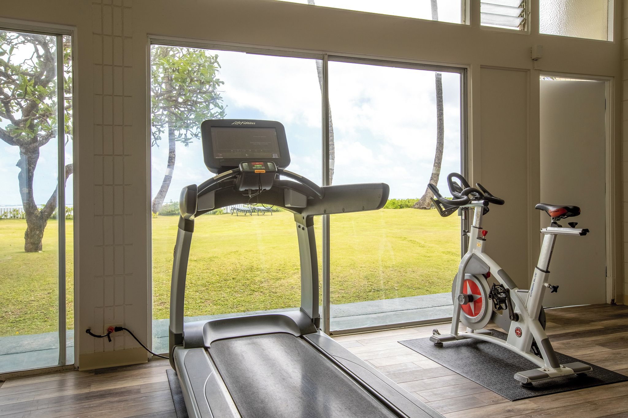 A home gym with a treadmill and an exercise bike, large windows with a view of a lawn and trees.