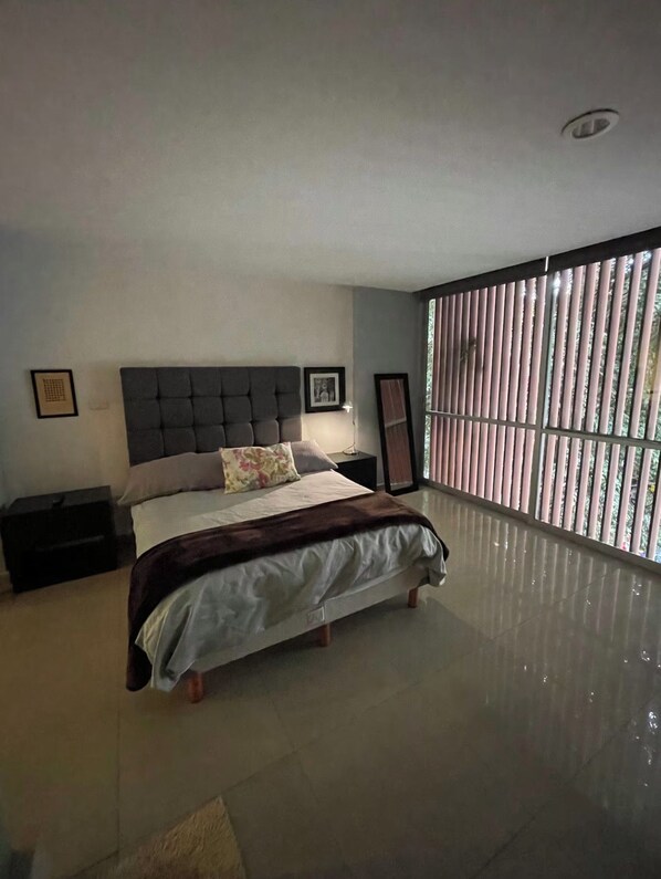 Ein Schlafzimmer mit einem großen Bett, einem Nachttisch, einer Fensterbank und einem Fenster mit Jalousien.