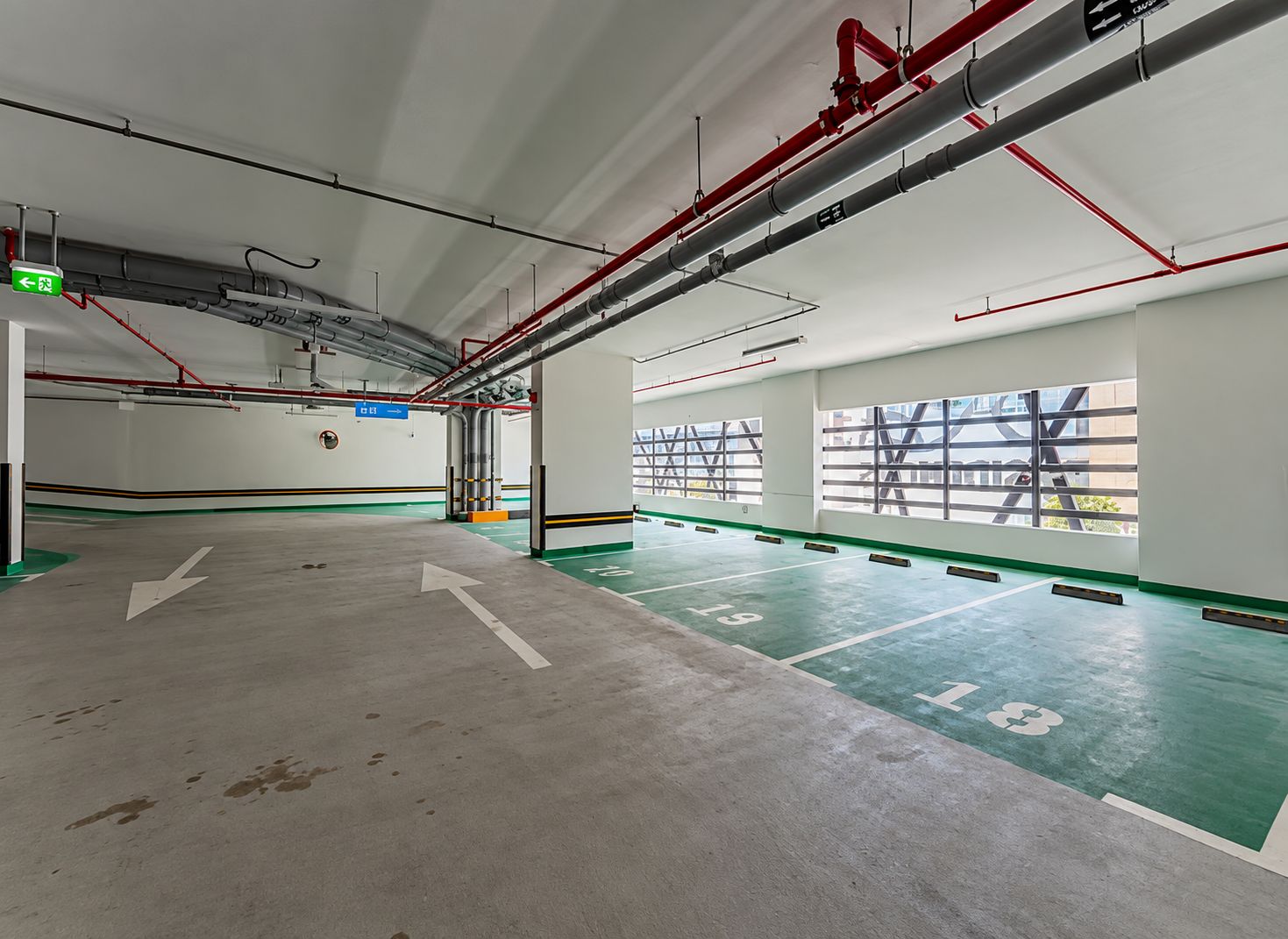 Un área de estacionamiento interior espaciosa, con flechas direccionales en el suelo, un letrero de salida verde y vistas al exterior a través de amplias ventanas.