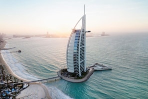Un hotel costero con un edificio de diseño en forma de vela, un paseo marítimo y un muelle que se adentra en el mar.