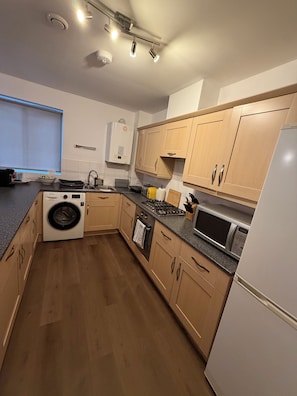 A compact kitchen with wooden cabinets, a built-in washing machine, a microwave, and a gas stove. - Zana's Apartment/6 beds / Near Train Station and Town Centre (Ipswich)