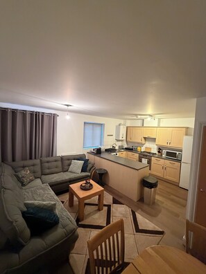 A modern living room with a sofa, coffee table, and a dining area. A kitchen with wooden cabinets and appliances is visible in the background. - Zana's Apartment/6 beds / Near Train Station and Town Centre (Ipswich)