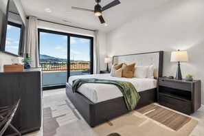 A modern hotel room with a large bed, a desk, a chair, a nightstand, a lamp, and a view of the mountains. - Waterpark View, Steps Away, Sauna + Arcade (Hurricane)