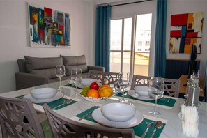 A dining area with a table set for four, including plates, bowls, and cutlery. There are chairs around the table, and a fruit bowl with oranges and apples. The room has a sofa and a painting on the wall. - Beachside • Balcony • Estoril Beach • Best Beach (Sal Rei)