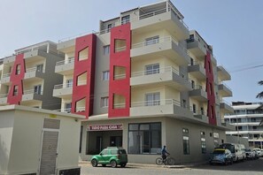 Un edificio residencial de varios pisos con balcones, un coche verde estacionado al frente y una persona en bicicleta.