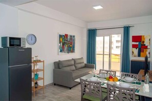 A compact living space with a kitchenette, a sofa, a dining table set for two, and a large window with curtains. - Beachside • Balcony • Estoril Beach • Best Beach (Sal Rei)