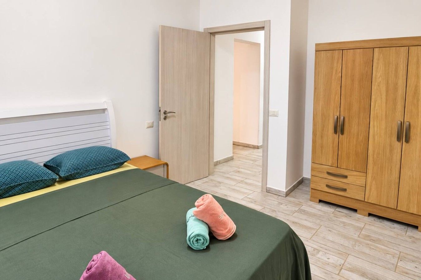 Een slaapkamer met een bed, kussens, een klein tafeltje, een kledingkast en een deur naar een andere kamer.