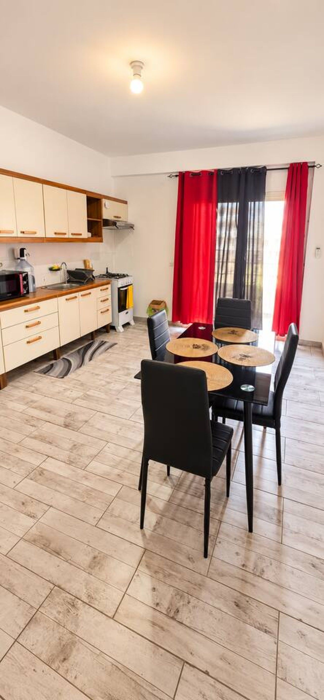 Une cuisine moderne avec des meubles en bois, un coin repas comprenant une table et des chaises, et un rideau rouge.