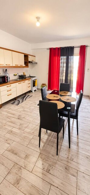 A modern kitchen with wooden cabinets, a dining area with a table and chairs, and a red curtain. - Beachside 2BR Apt • Balcony • Estoril beach (Sal Rei)