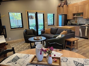 A modern living room with a grey sofa, a coffee table, and a dining table with a tray. There are windows with a view of a wooden deck and trees. - Mountain-modern vacation rental with Full Kitchen, King Bed, Fireplace, Hot Tub (Winthrop)