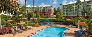 A hotel pool area with lounge chairs and umbrellas, surrounded by greenery and a multi-story building with balconies. - Theme park capital of the world! 2 bedroom, two bath, peaceful retreat (Orlando)
