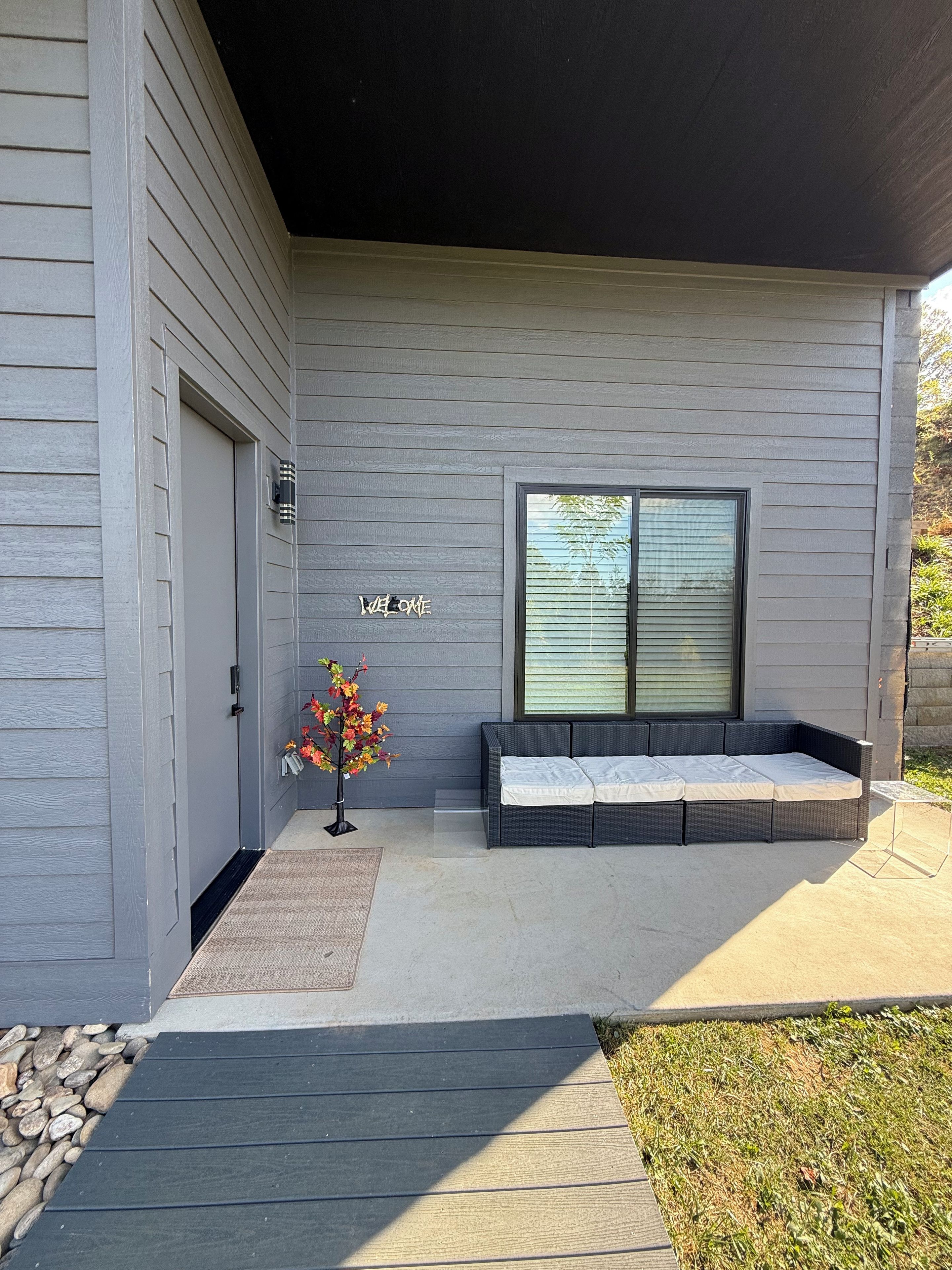 A modern house with a grey exterior, a welcome sign, a covered patio with a sofa, and a small tree with autumn leaves.
