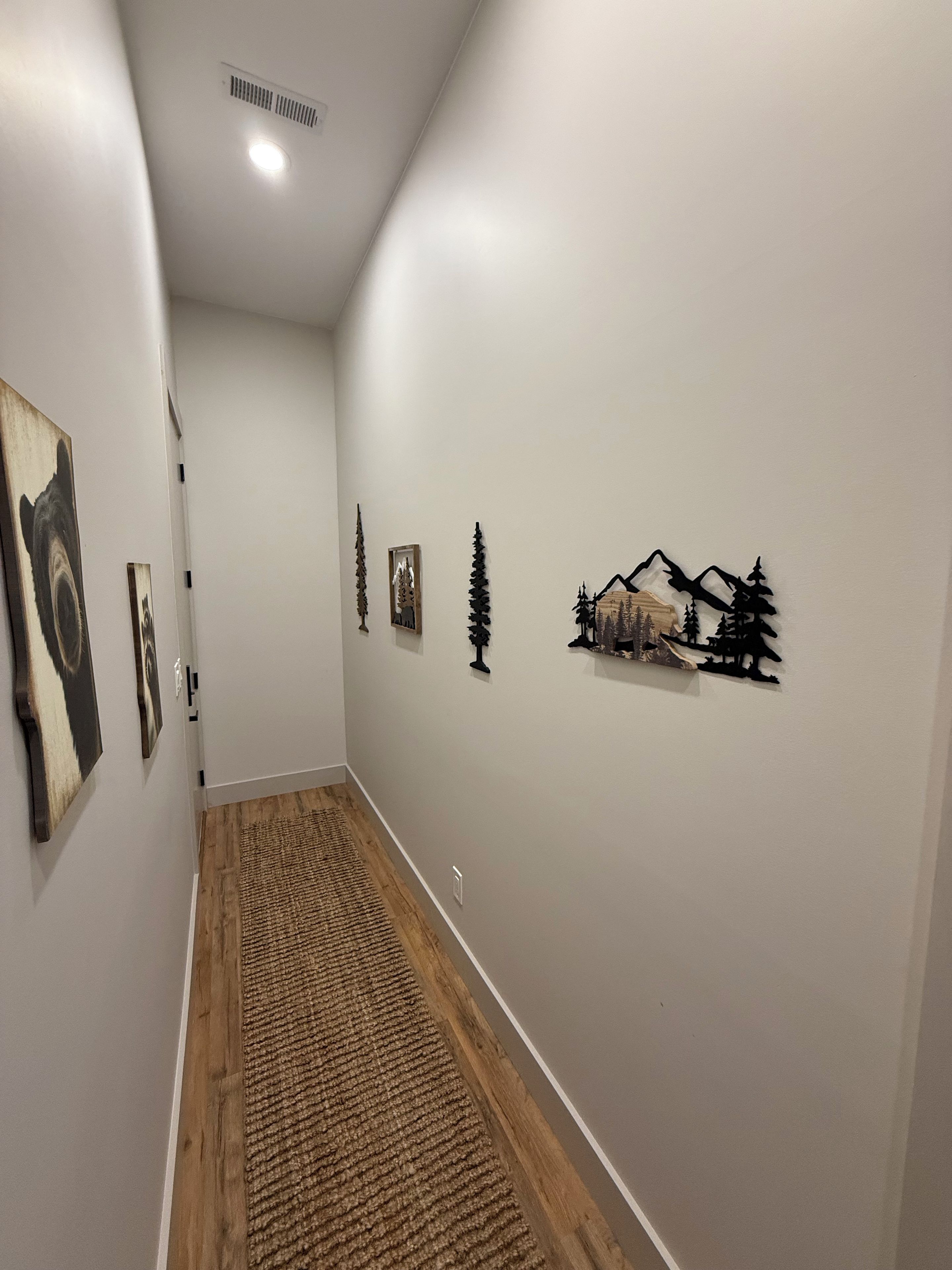 Un pasillo con suelo de madera, paredes blancas y diversas decoraciones en la pared, incluyendo un paisaje de montaña y árboles.