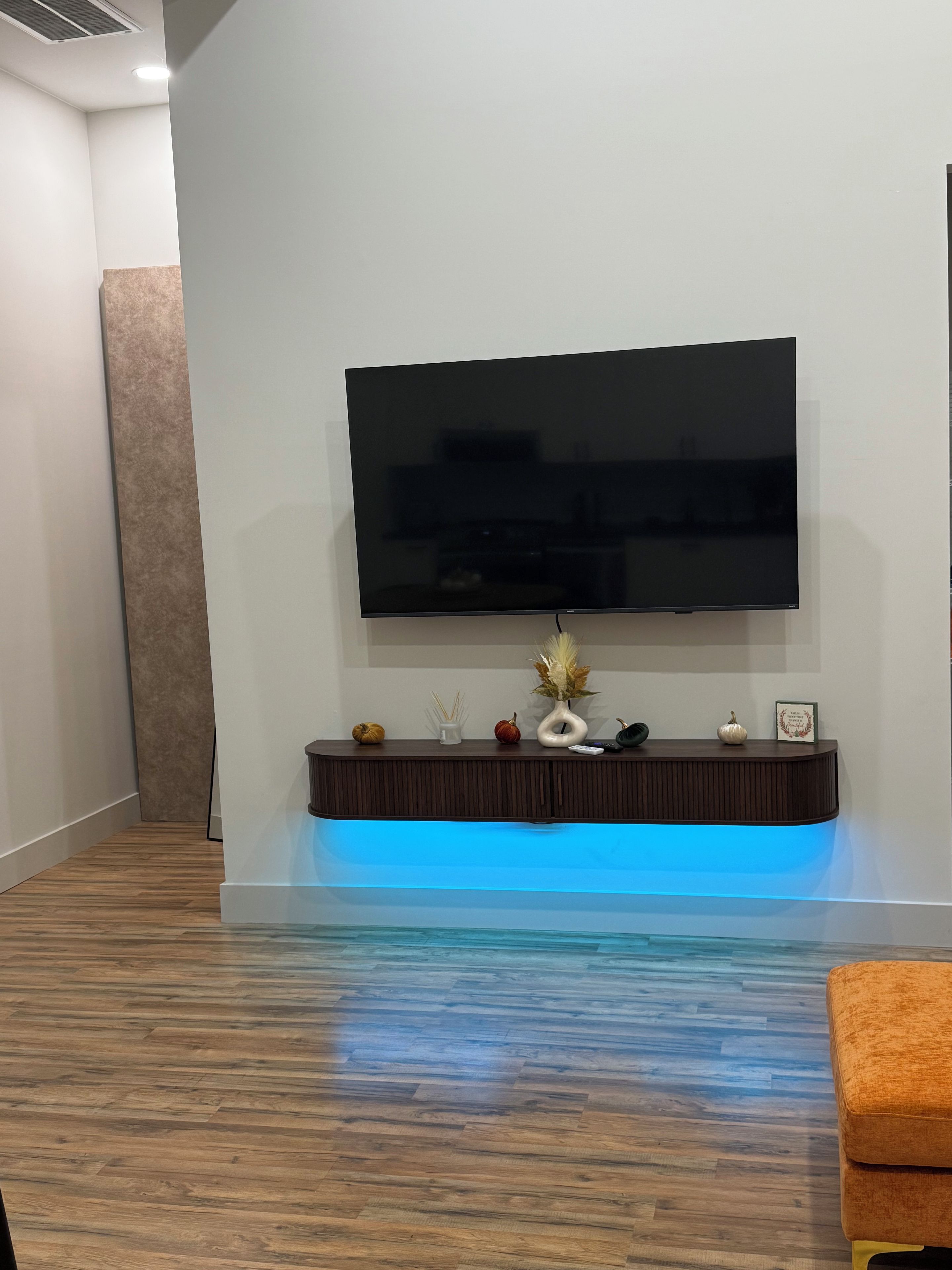 Een moderne woonkamer met een flatscreen televisie aan de muur, een houten vloer en een blauw verlichte console met decoratieve objecten.