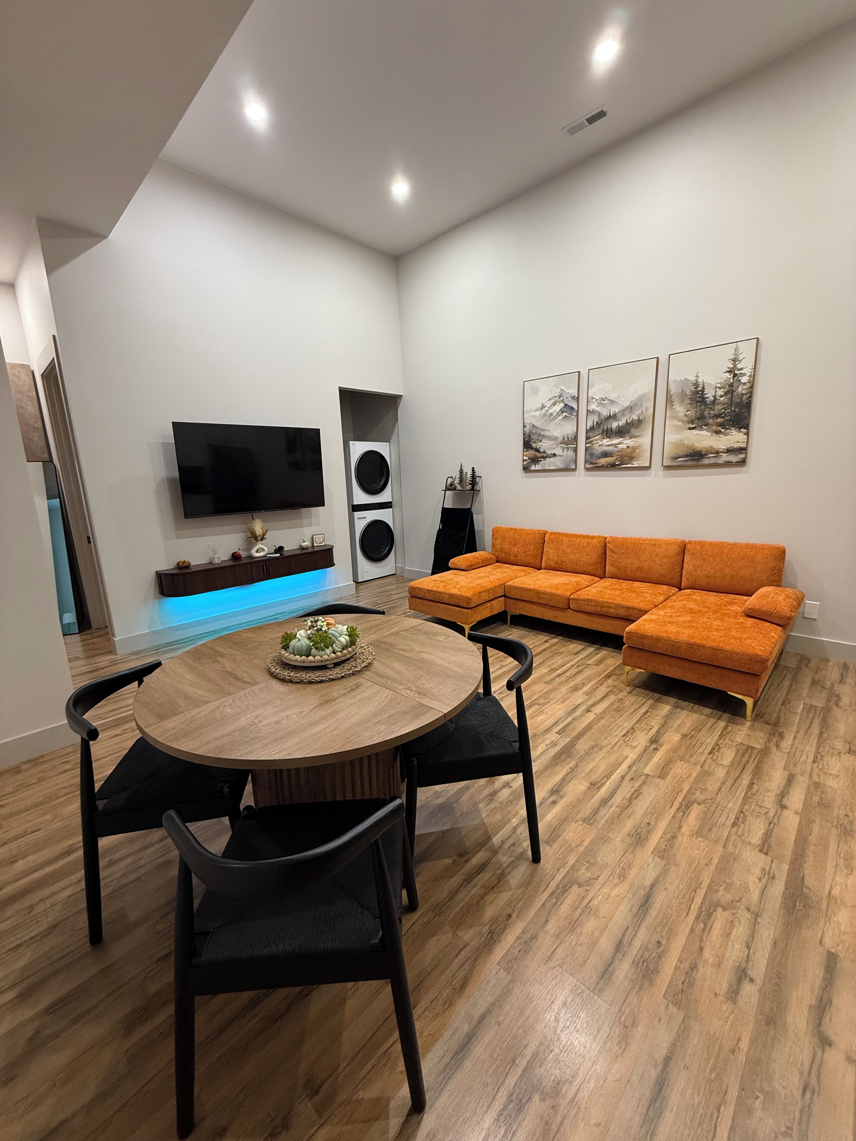 A modern living room with a round wooden dining table, black chairs, an orange sectional sofa, a flat-screen TV, and a wall-mounted speaker system.