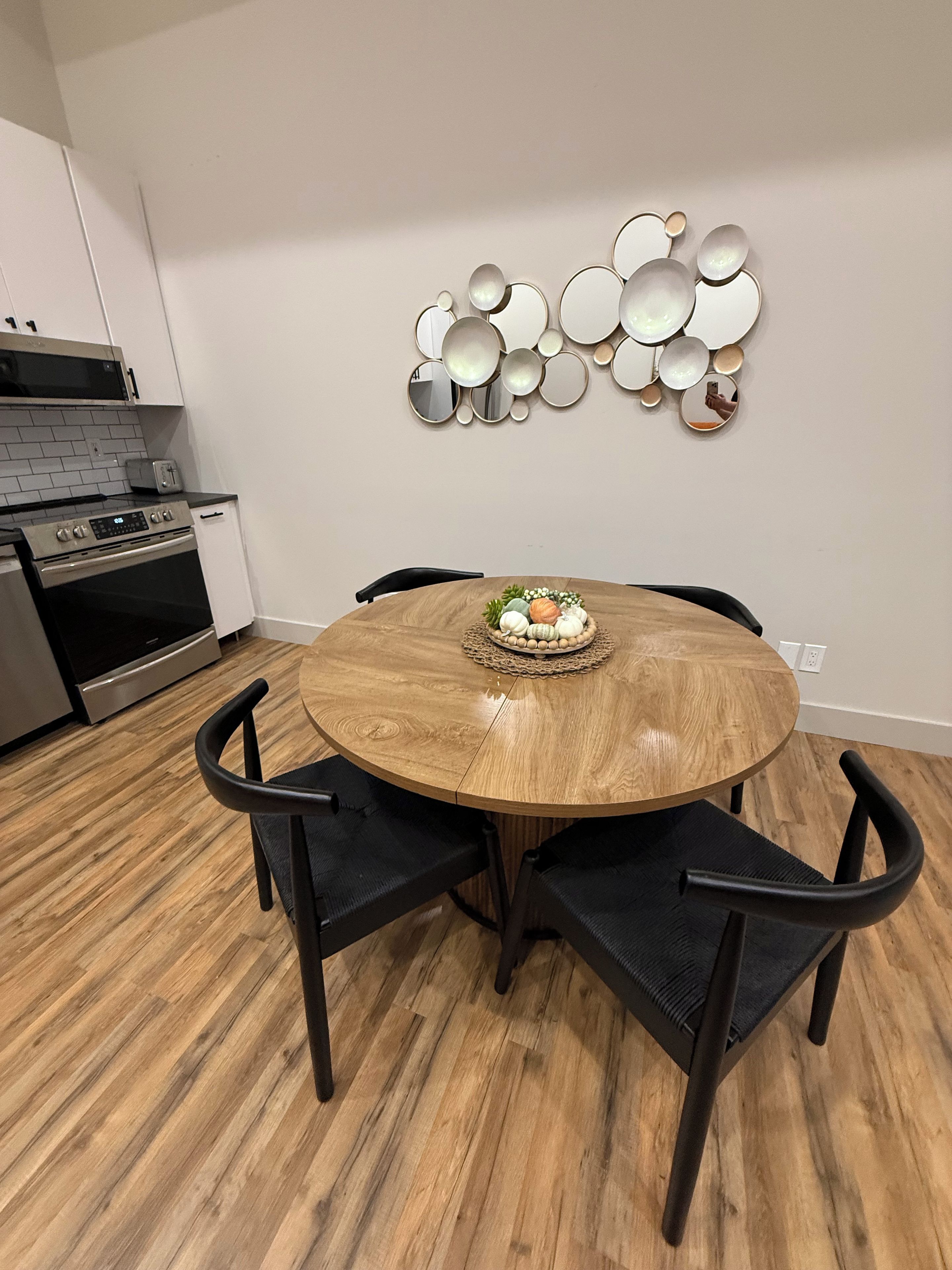 A dining area with a round wooden table, four black chairs, and a decorative wall piece above the table.