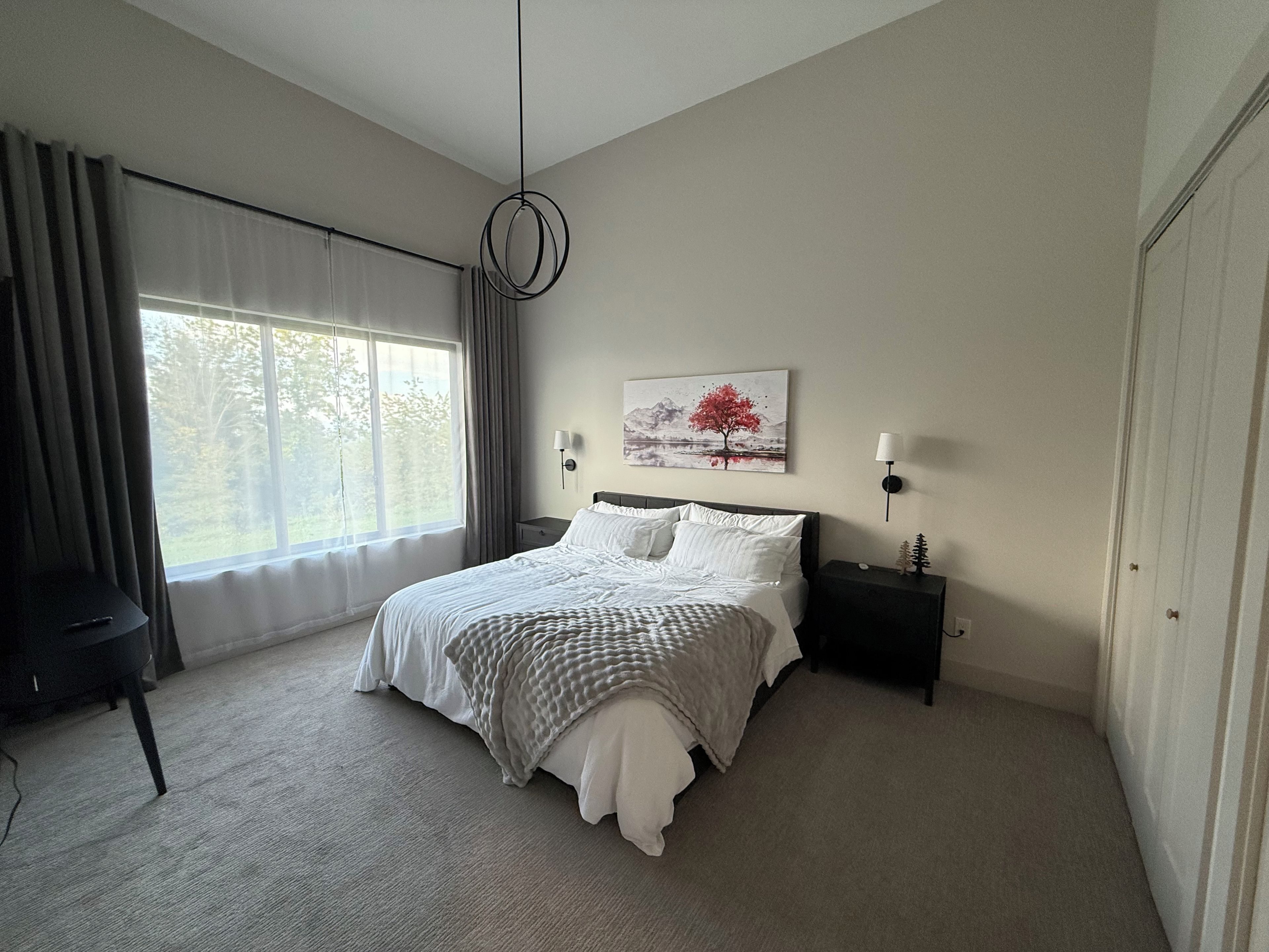 Une chambre à coucher avec un grand lit, une table de chevet, une lampe, un tableau au mur et une fenêtre avec des rideaux.