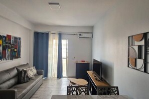 Een moderne woonkamer met een bank, een eettafel en een televisie. Er is een raam met gordijnen en een airconditioning op de muur.