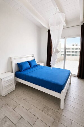 A bedroom with a bed, a nightstand, a window with curtains, and a view of buildings outside. - 2 apartments for up to 8 guests in Estoril beach (Sal Rei)