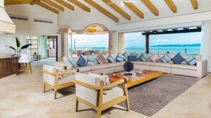 Un vaste espace de vie comprenant un grand canapé, des chaises en bois et une table basse. Un balcon avec vue sur la mer est visible à travers de grandes fenêtres.