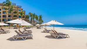 Ein Strand mit Liegestühlen und Sonnenschirmen, Palmen und einem Hotel im Hintergrund.