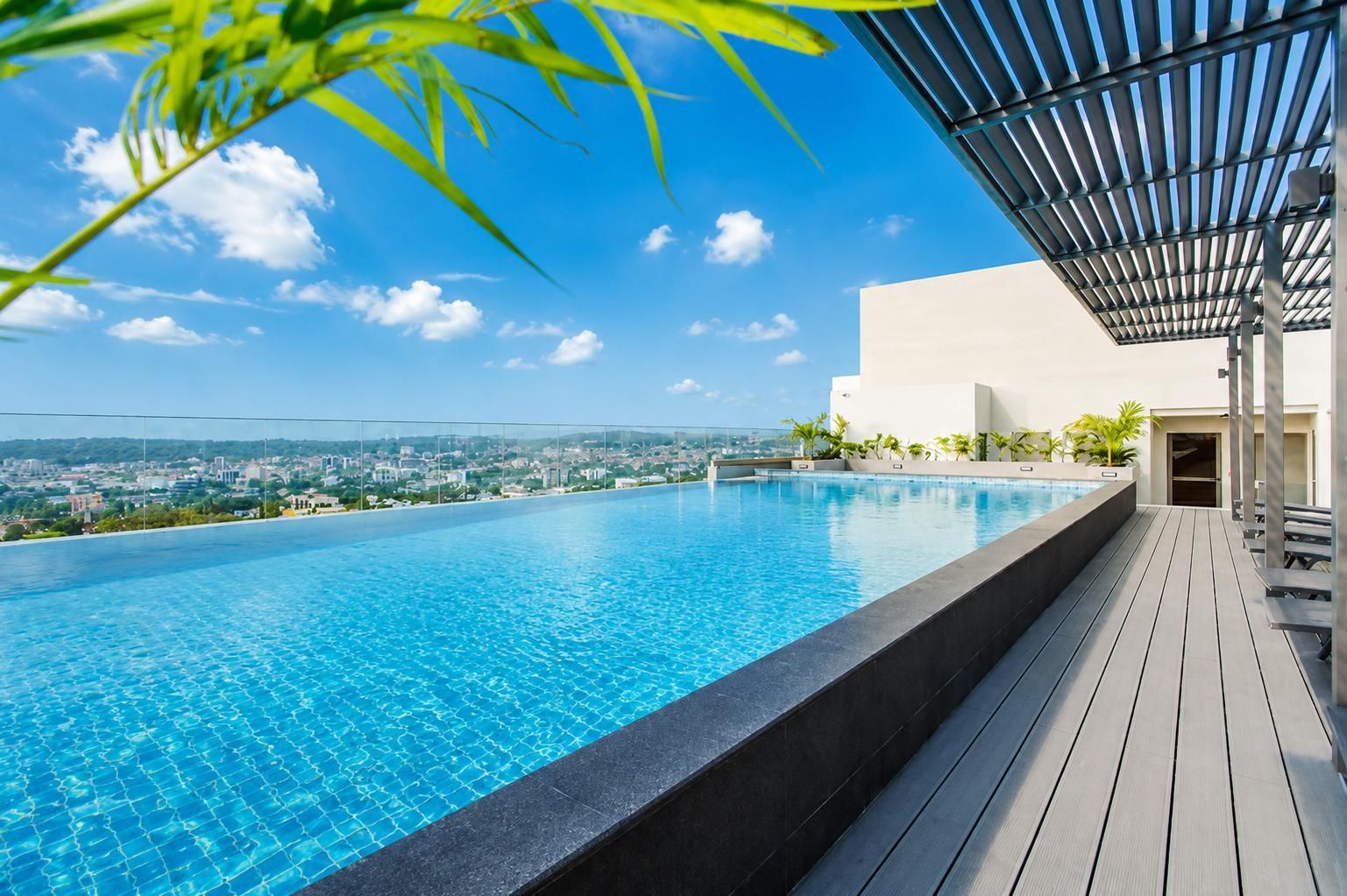 Une piscine à débordement située sur le toit, offrant une vue sur la ville, avec une terrasse en bois et un espace salon couvert.