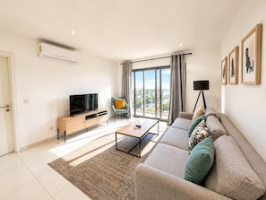 Una sala de estar moderna con un sofá, televisor, mesa de centro y vistas a la ciudad a través de amplias ventanas.