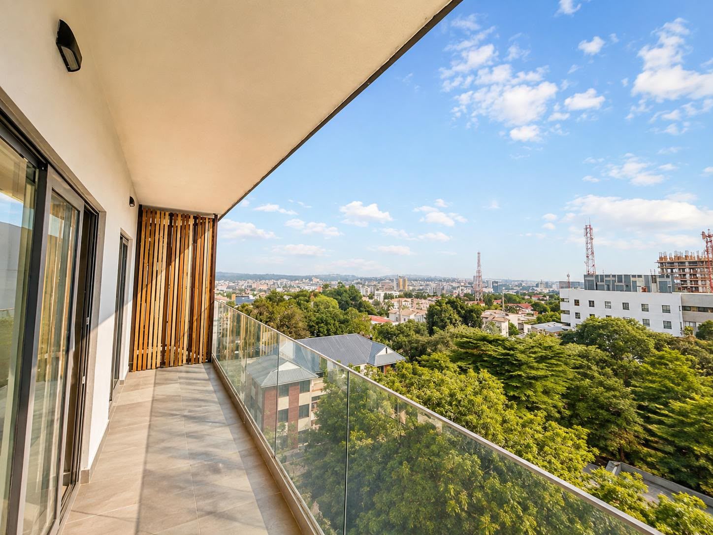 Un balcon doté d’une balustrade en verre, offrant une vue sur un paysage urbain avec des immeubles et des arbres.