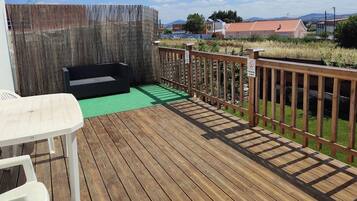 Una terraza de madera con una mesa de picnic y un sofá negro, rodeada por una valla de madera y una zona ajardinada.