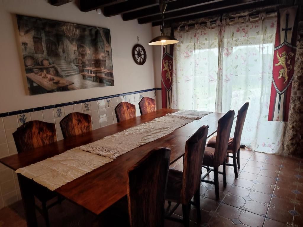 Un comedor con una larga mesa de madera, seis sillas, un cuadro decorativo en la pared, una araña y una ventana con cortinas.