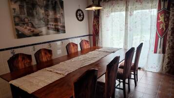 Un comedor con una larga mesa de madera, seis sillas, un cuadro decorativo en la pared, una araña y una ventana con cortinas.