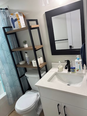 A bathroom with a white toilet, a shelf with toiletries, a mirror, and a shower curtain. - Cozy Basement Suite - Home Away From Home (Saskatoon)