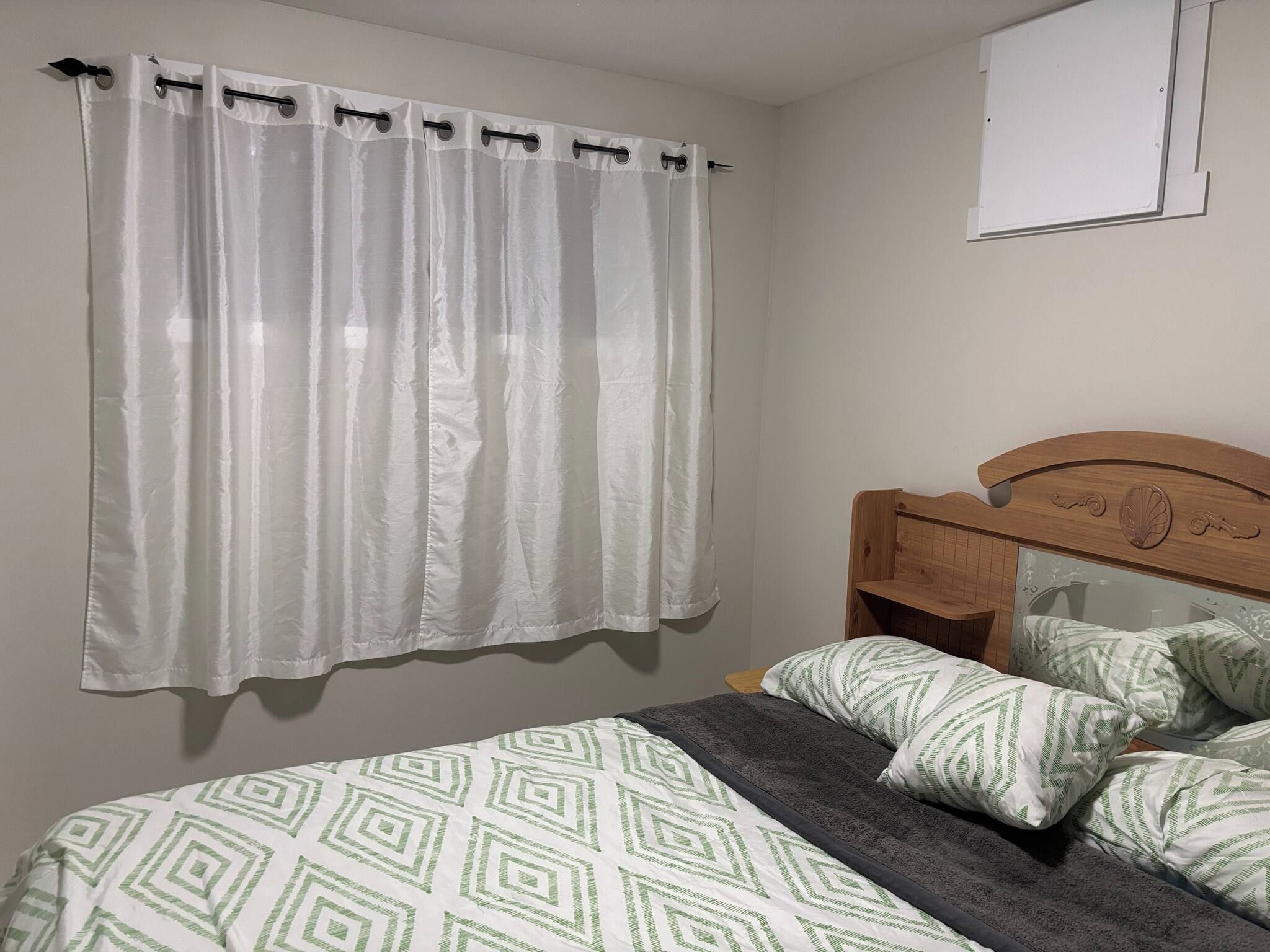 A bedroom with a bed, pillows, a wooden headboard, and a white curtain.