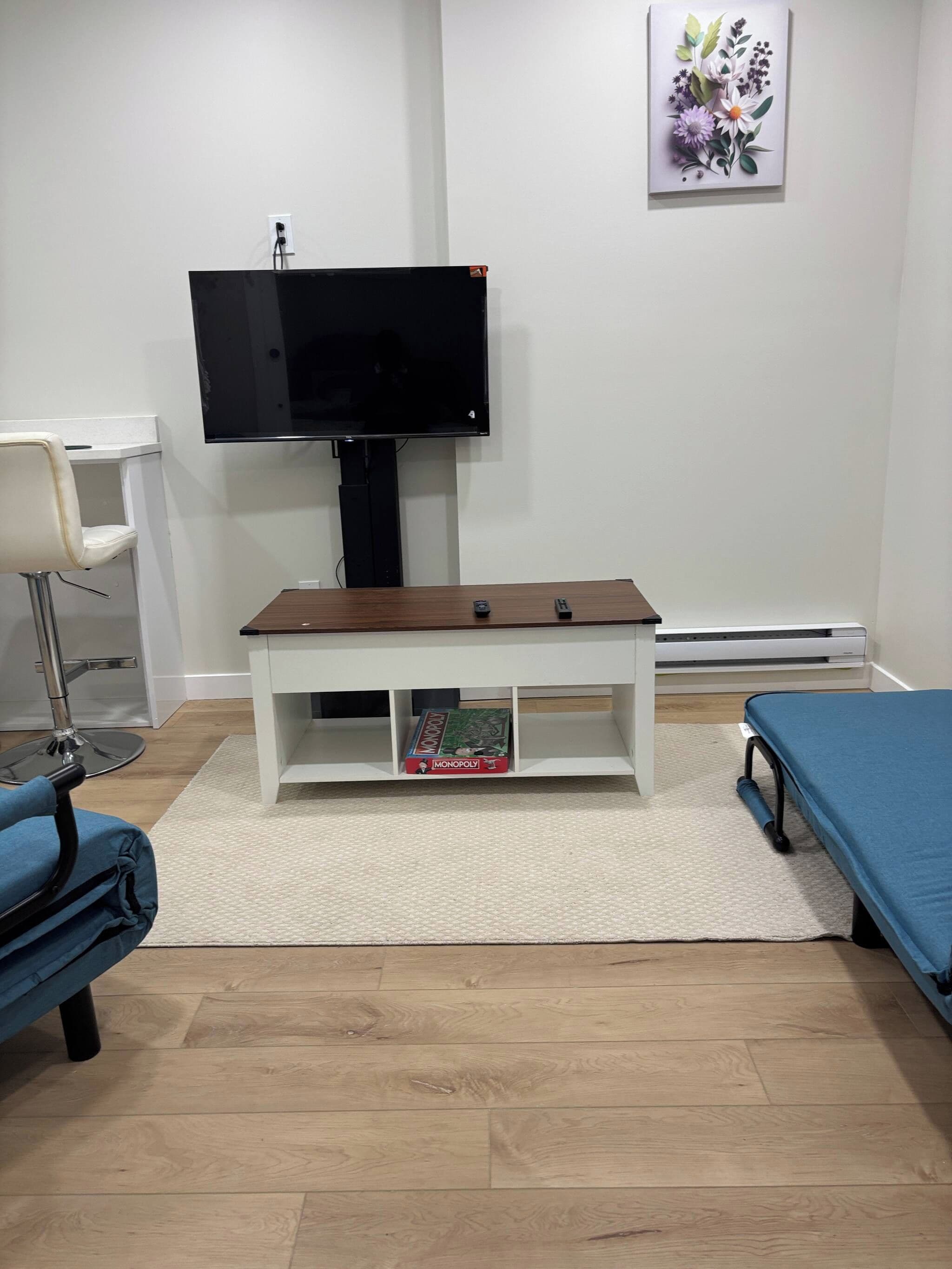 A room with a television mounted on the wall, a white desk with a chair, a blue bed, and a game board on the floor.