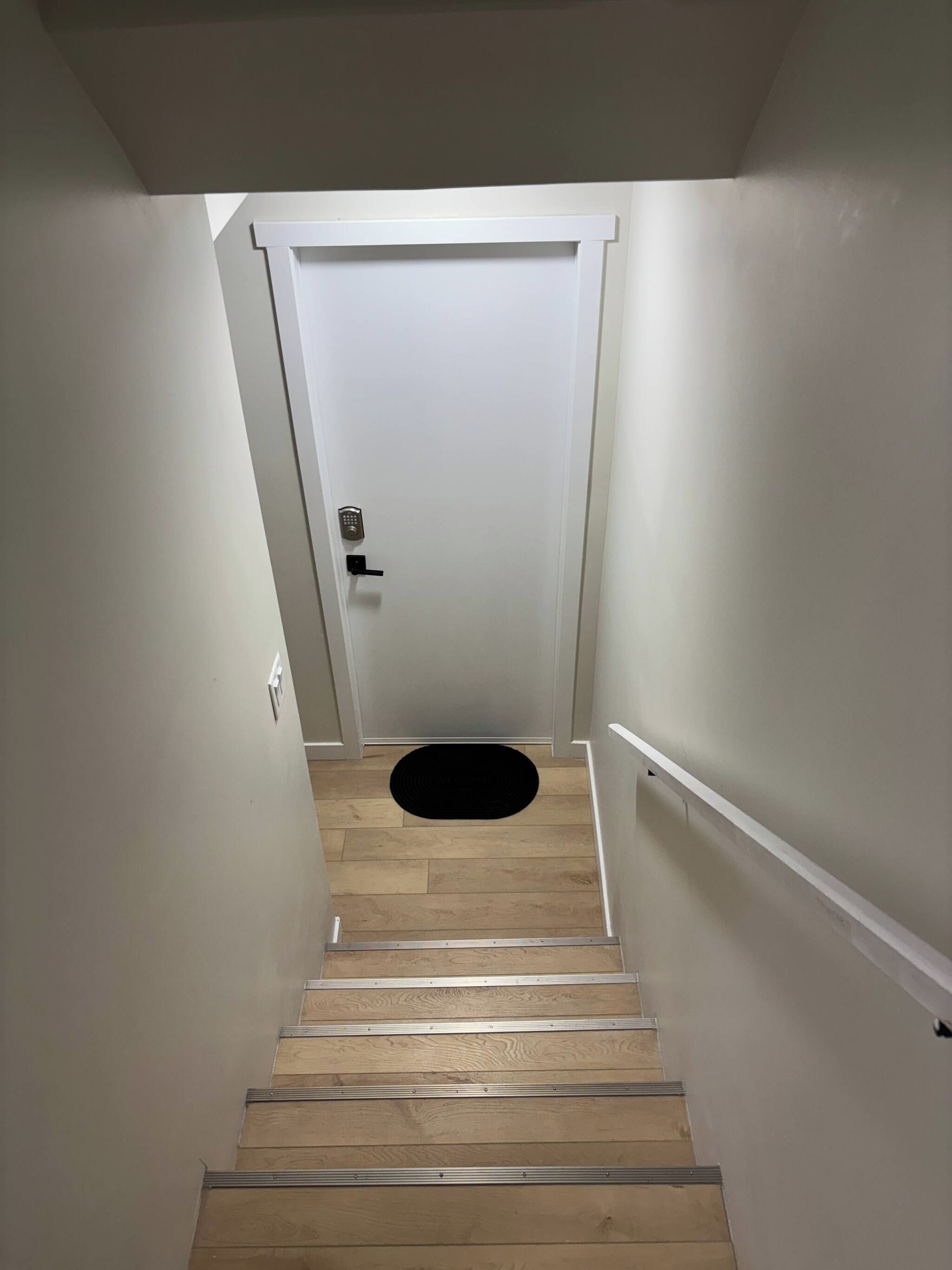 A staircase with a black doormat at the bottom, leading to a white door with a keypad lock.