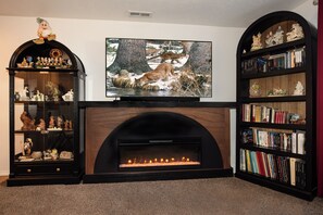A living room with a fireplace, a television, and two bookshelves filled with books and decorative items. - Location, Location, Location (Sandy)