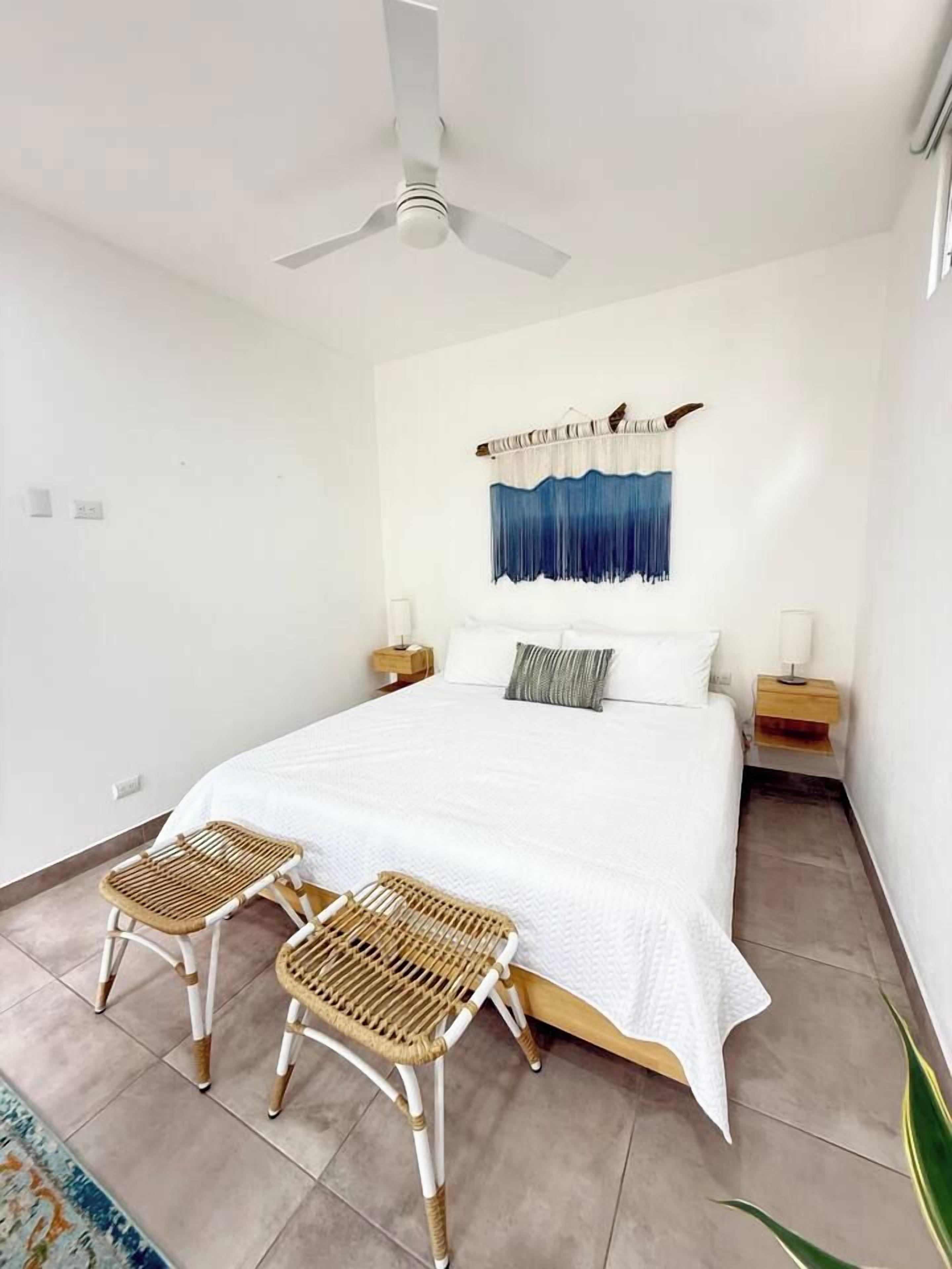 A bedroom with a bed, two nightstands, a ceiling fan, a wall-mounted tapestry, and two wicker chairs.