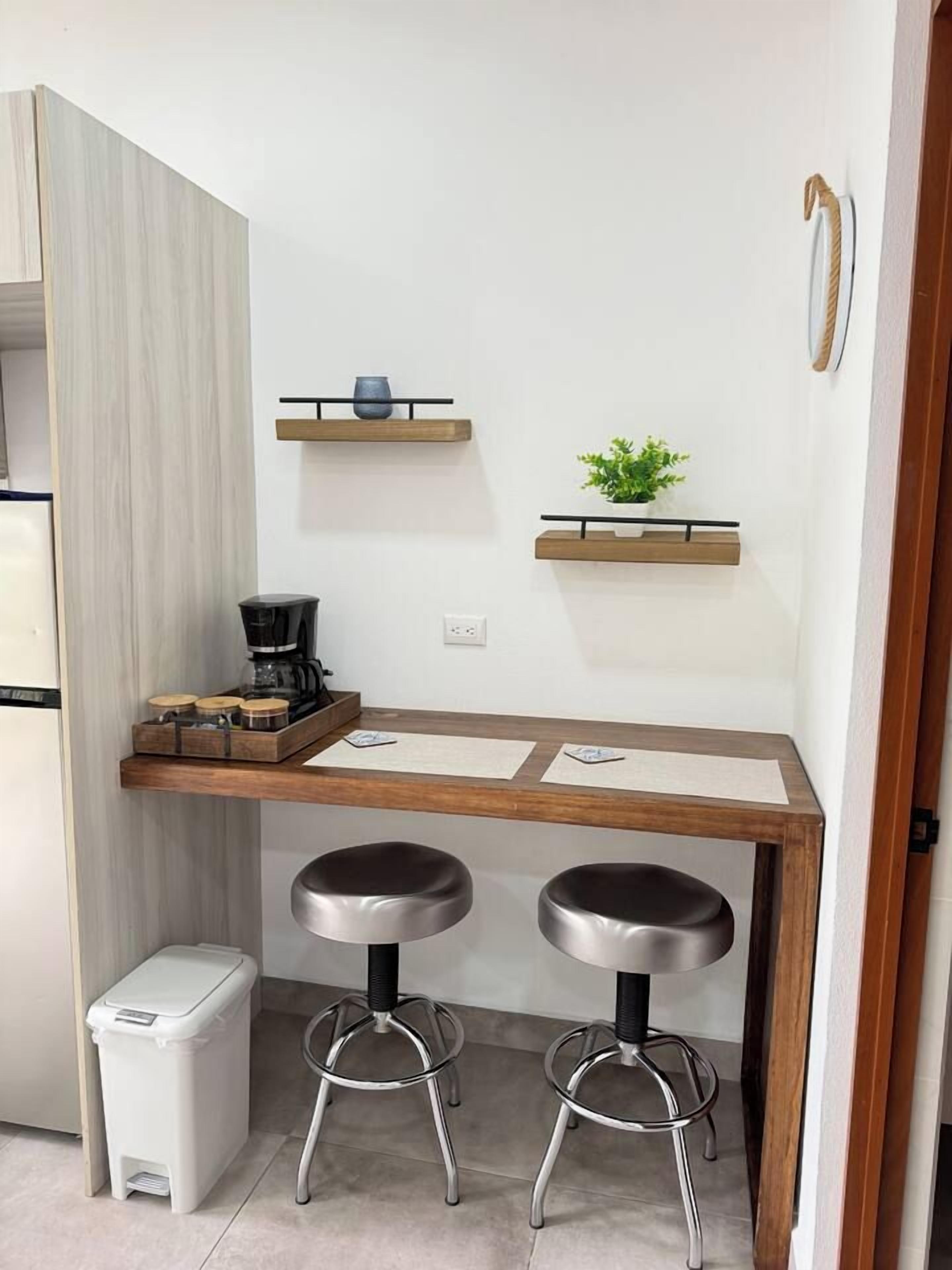A compact kitchenette with a coffee maker, two stools, and a small trash bin.
