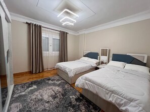 Ein Hotelzimmer mit zwei Betten, einem großen Fenster mit Vorhängen, einer modernen Deckenleuchte und einem gemusterten Teppich.