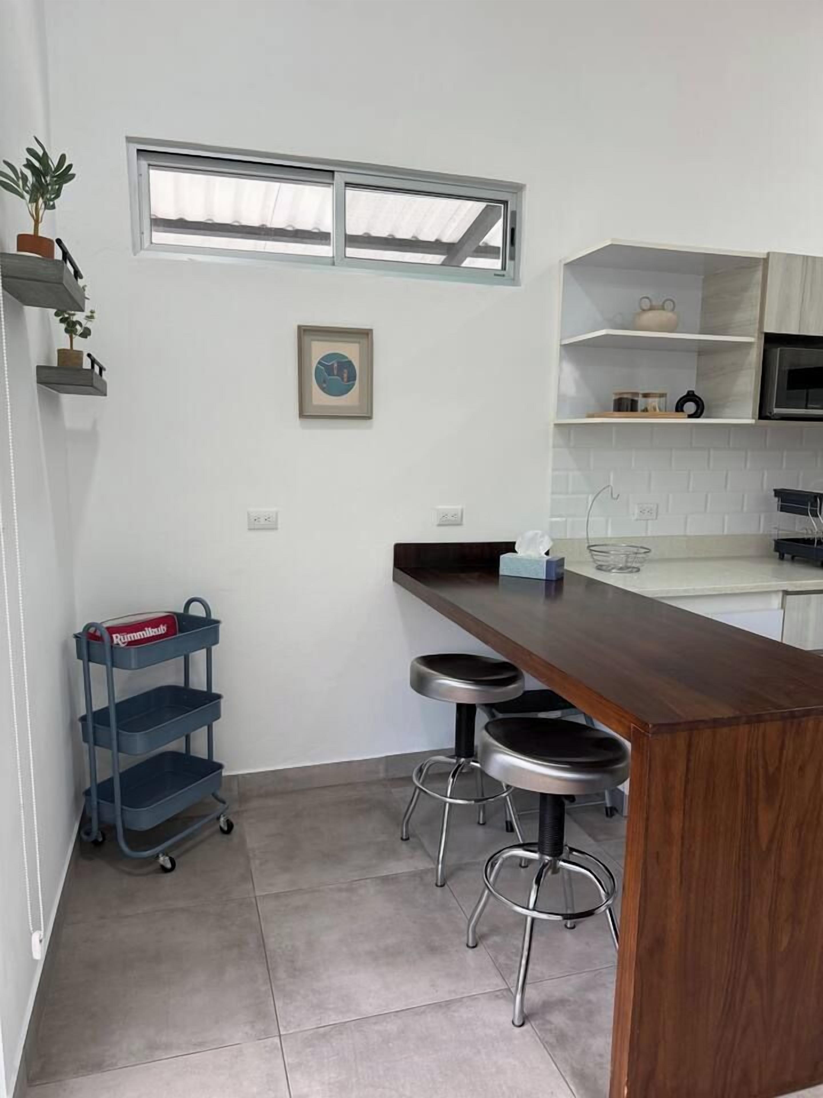 A compact kitchen with a wooden countertop, two stools, a small shelf unit, and a potted plant.