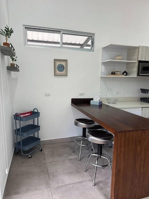 A compact kitchen with a wooden countertop, two stools, a small shelf unit, and a potted plant. - Casa Swell at Costa Lofts (Jaco)