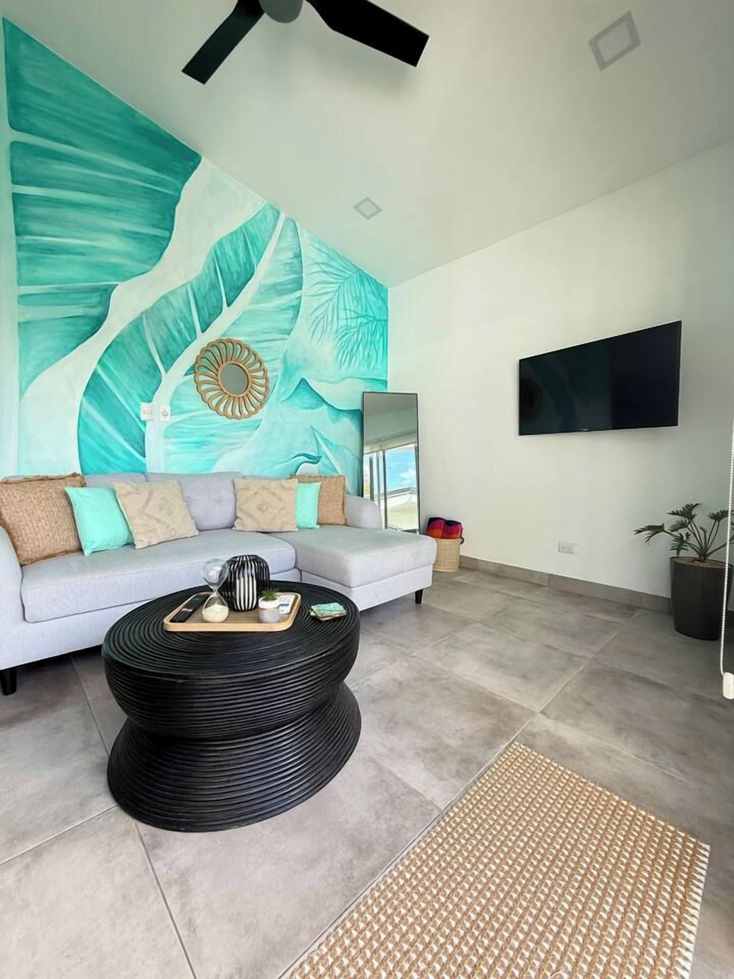 A living room with a sofa, a round black coffee table, a flat-screen TV mounted on the wall, and a large abstract mural on the wall.