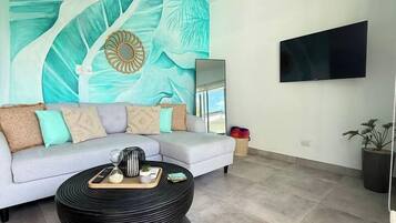 A living room with a sofa, a round black coffee table, a flat-screen TV mounted on the wall, and a large abstract mural on the wall.