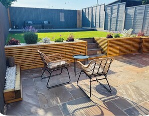 A patio with two wicker chairs and a small round table, surrounded by wooden planters with various plants. - COSY HOME - KINGSIZE BED & PRIVATE PARKING (Saughall Massie)